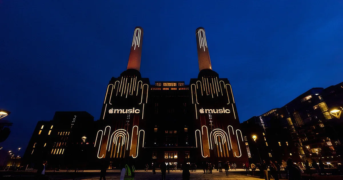 Battersea Power Station in London at dusk, its two front chimneys and facade lit with white arched light installations and the new lowercase Apple Music wordmark projected on each side.