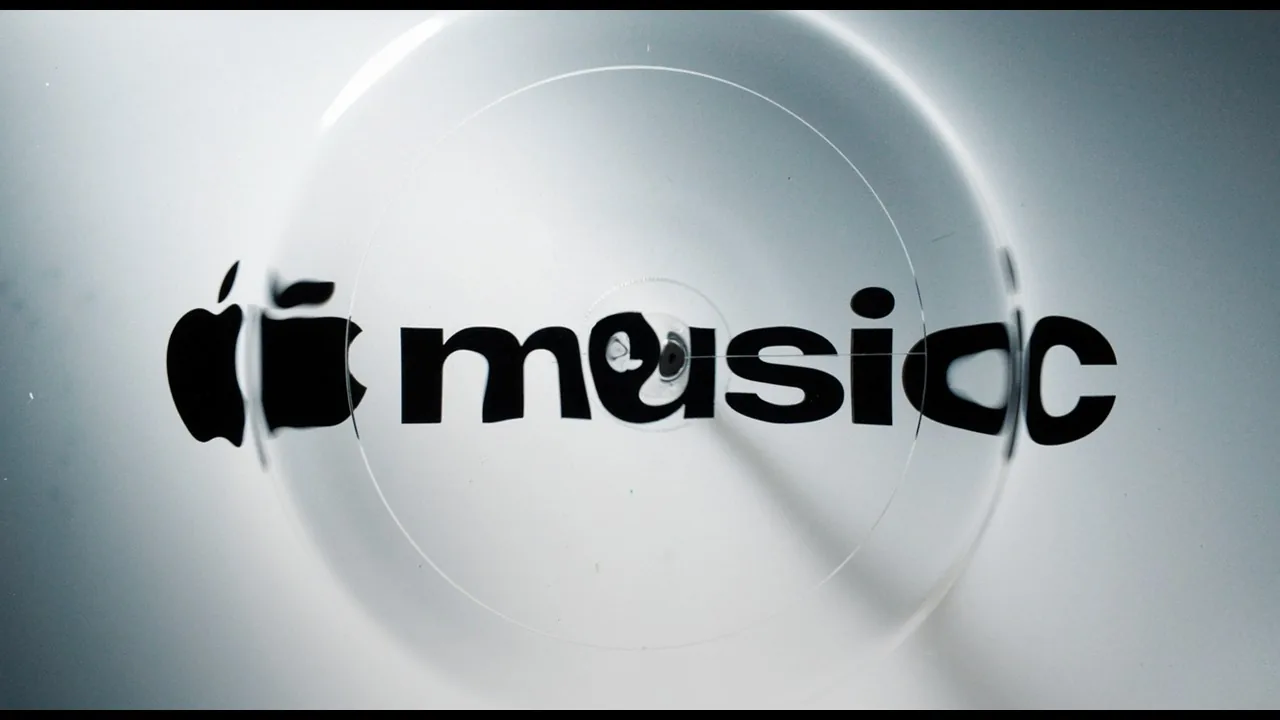The new Apple Music wordmark seen through a rippling layer of oil: the Apple symbol and the word music in bold lowercase, warped and refracted, with faint circular ripples spreading across a pale grey surface.
