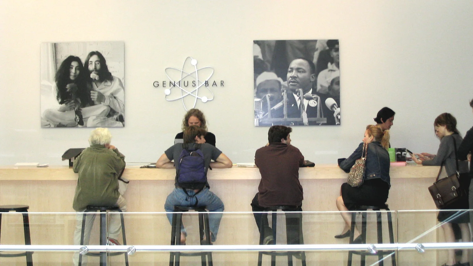 The Genius Bar at the Apple Store in SoHo, New York, in 2003: customers on stools at a long pale-wood counter, a Genius behind it, large black-and-white portraits of John Lennon and Yoko Ono and of Martin Luther King Jr. on the wall above.
