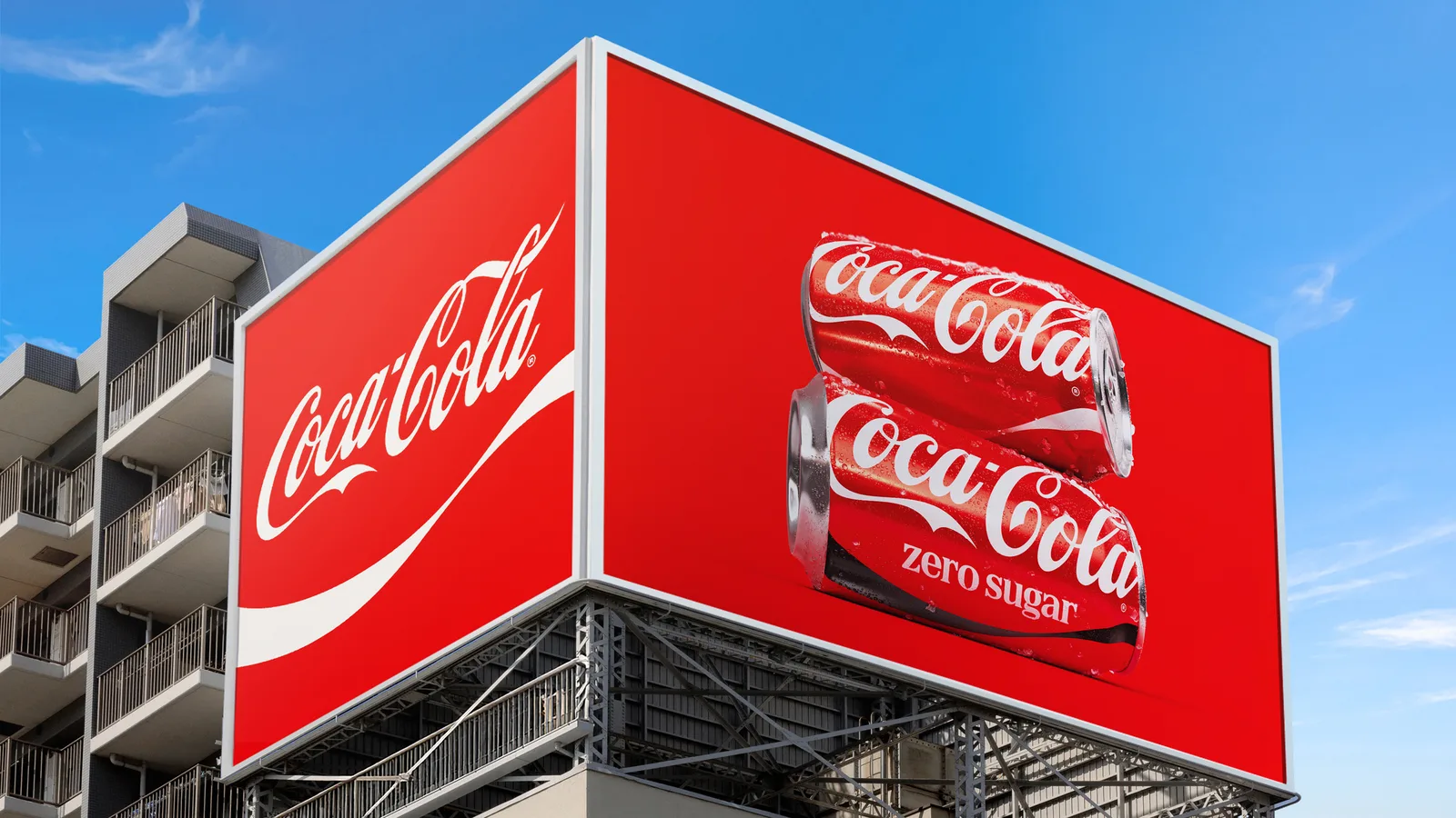 A large corner billboard on a city building, red with the white Coca-Cola script and ribbon on one face and two cans, Original Taste and Zero Sugar, on the other.